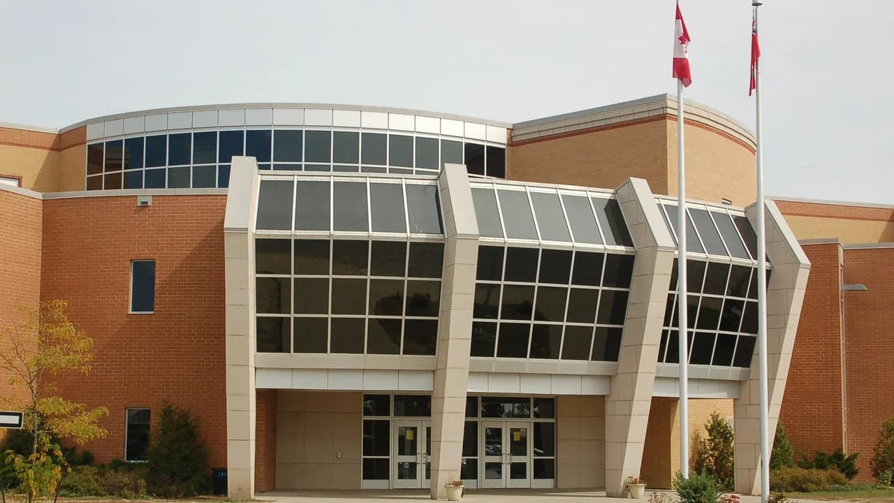 Entrance of All Saints Catholic Secondary School in Whitby, Ontario — a brick school building with a glass atrium entryway, Canadian and Ontario flags on flagpoles.