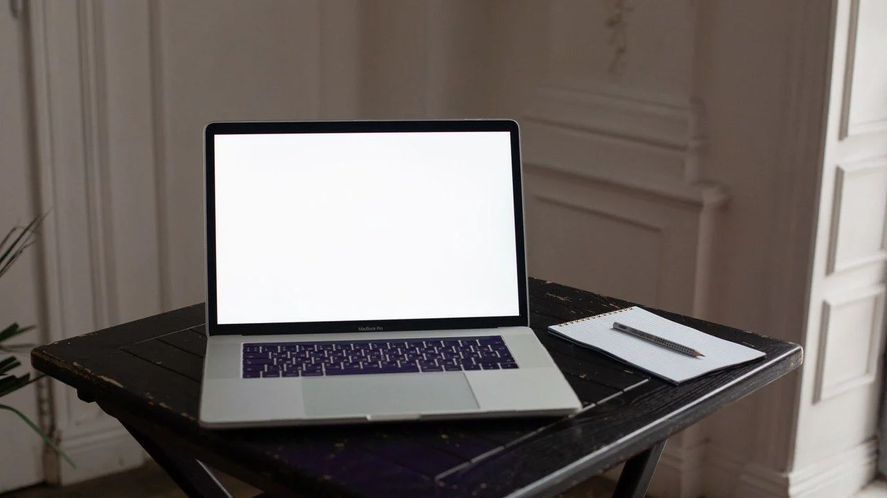 Laptop with a blank screen on a wooden table next to a notebook and pen; no people visible