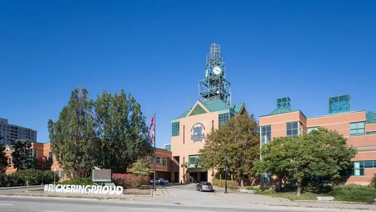 Pickering City Hall's brick civic building with its distinctive clock tower under a clear blue sky, with a #PICKERINGPROUD sign in the foreground.