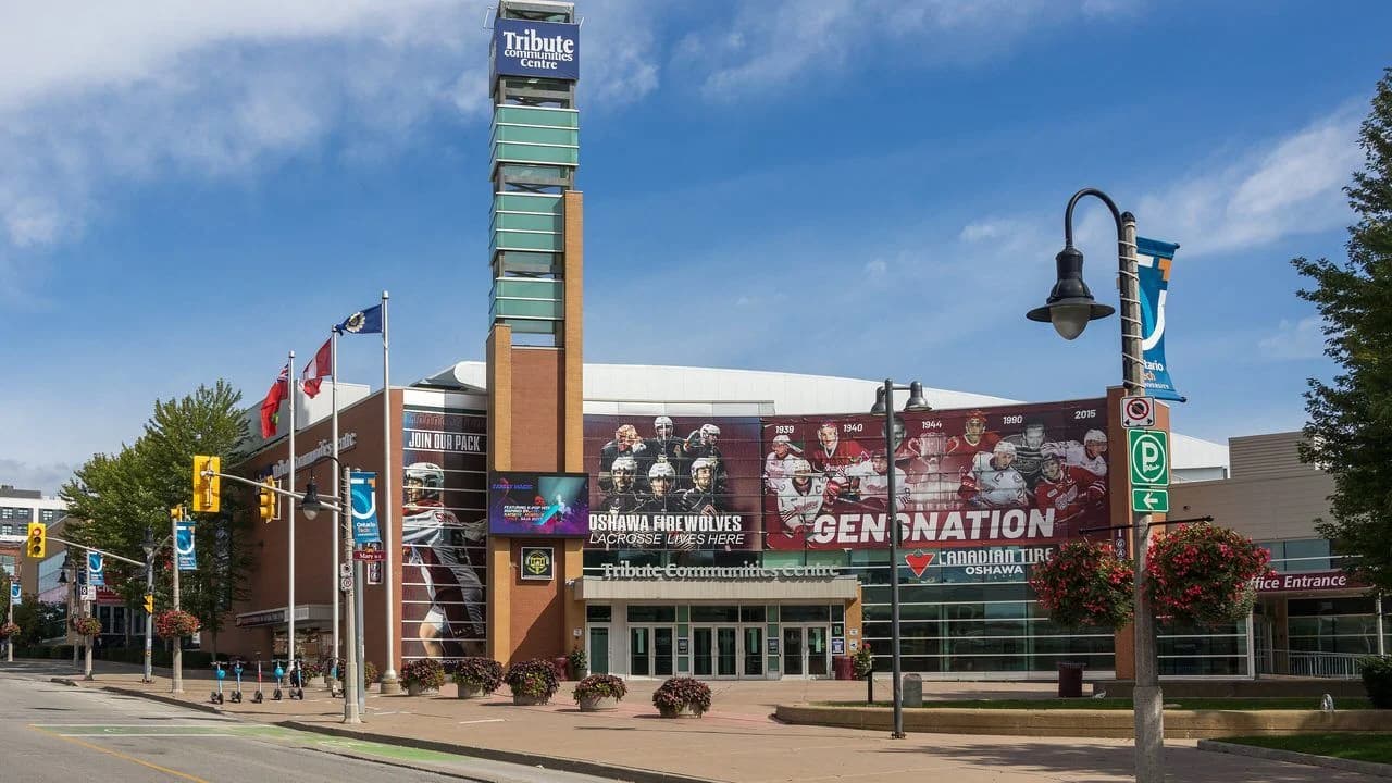 Exterior of the Tribute Communities Centre, home arena of the Oshawa Generals, on Athol Street in downtown Oshawa on a clear day; the brick facade carries large Oshawa Generals mural wraps, team flags fly out front, and the arena's tall tower sign rises above the entrance.