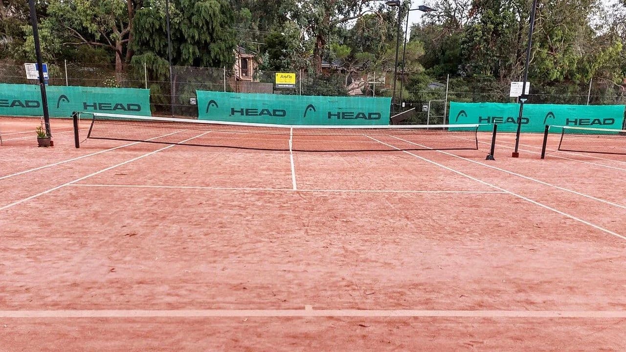 An empty clay tennis court with a net, green windscreens and trees in the background