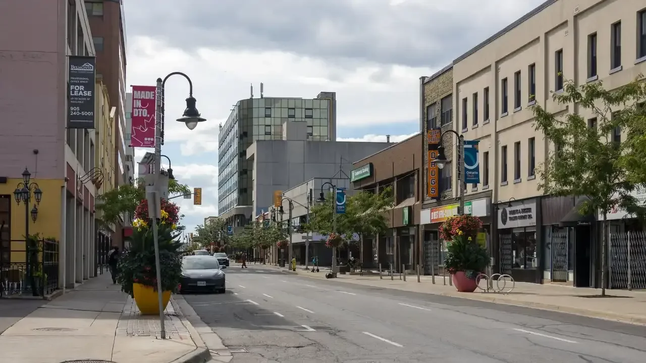 Downtown Oshawa's King Street lined with storefronts, banners, planters and parked cars under a cloudy sky.