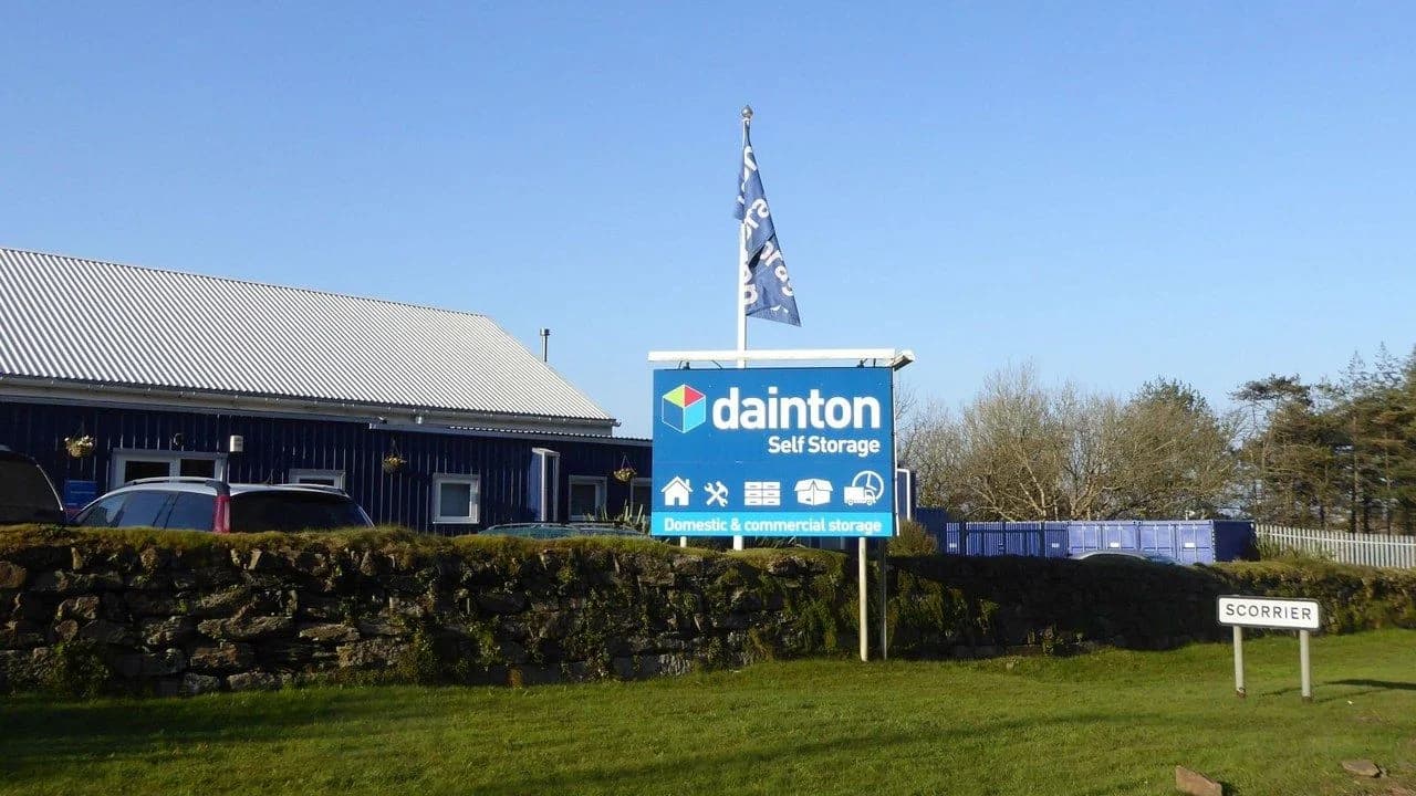 A self-storage facility exterior with rows of storage containers behind a stone wall and a blue 'self storage' sign; no people visible.